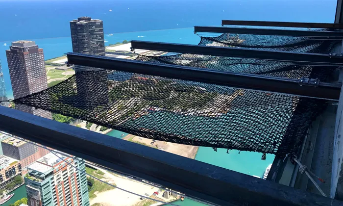 Black safety netting stretched between steel outriggers on a high-rise overlooking a harbor and city skyline